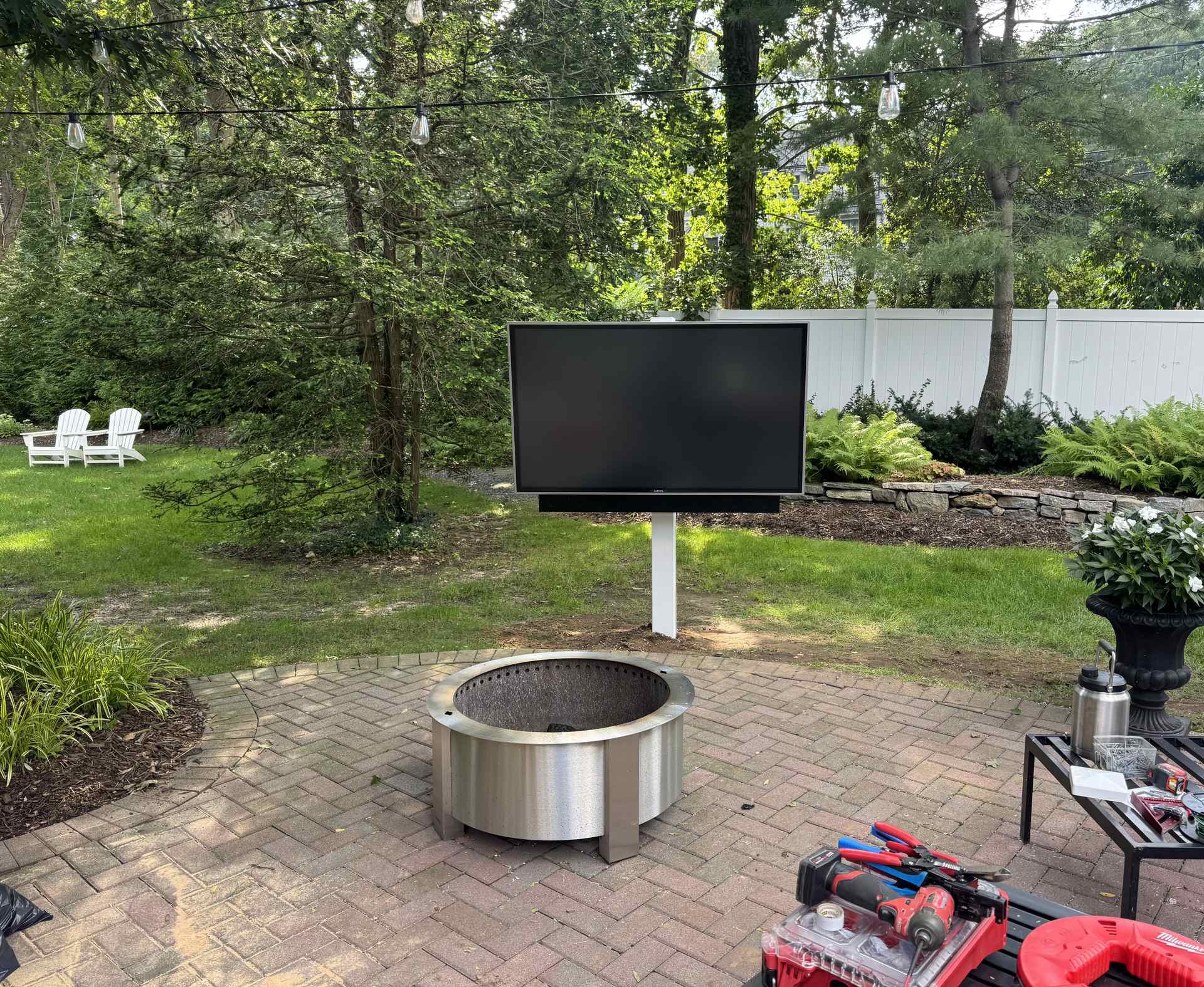 Outdoor TV mounted on a weatherproof post near a stainless steel fire pit on a wooded Northport, NY patio